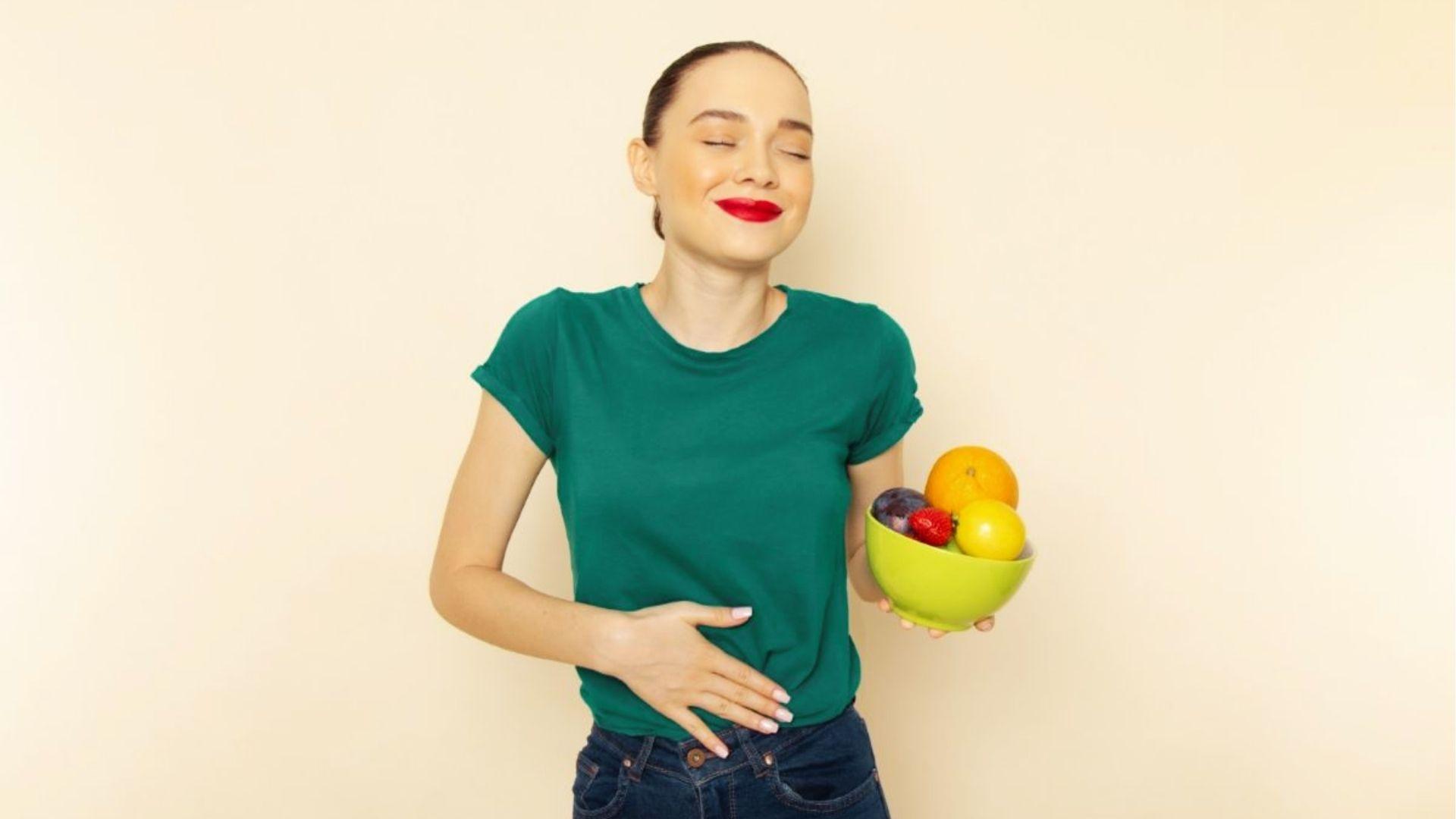 Smiling woman in green shirt holding bowl of fruit while touching her stomach against beige background