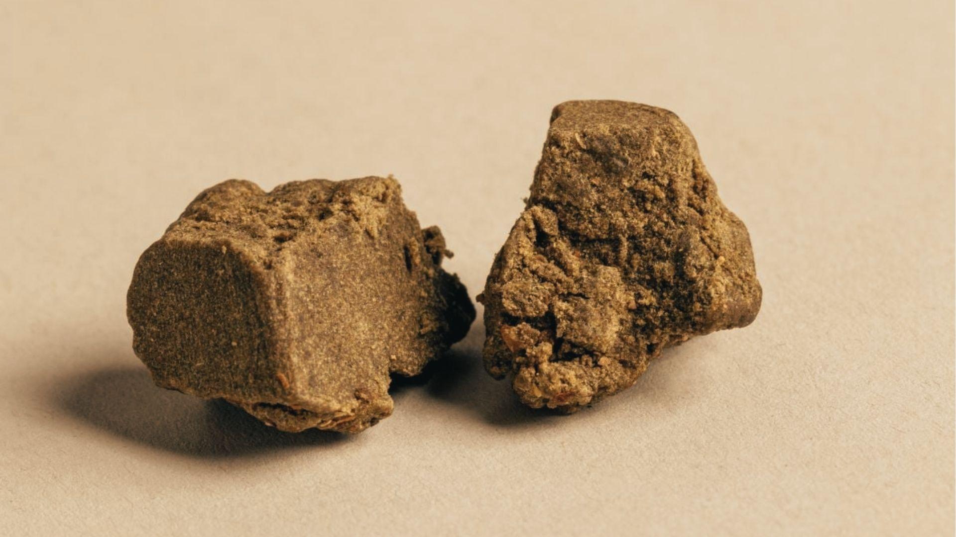 Two rough brown chunks of textured hashish resin displayed on neutral beige paper with soft shadows