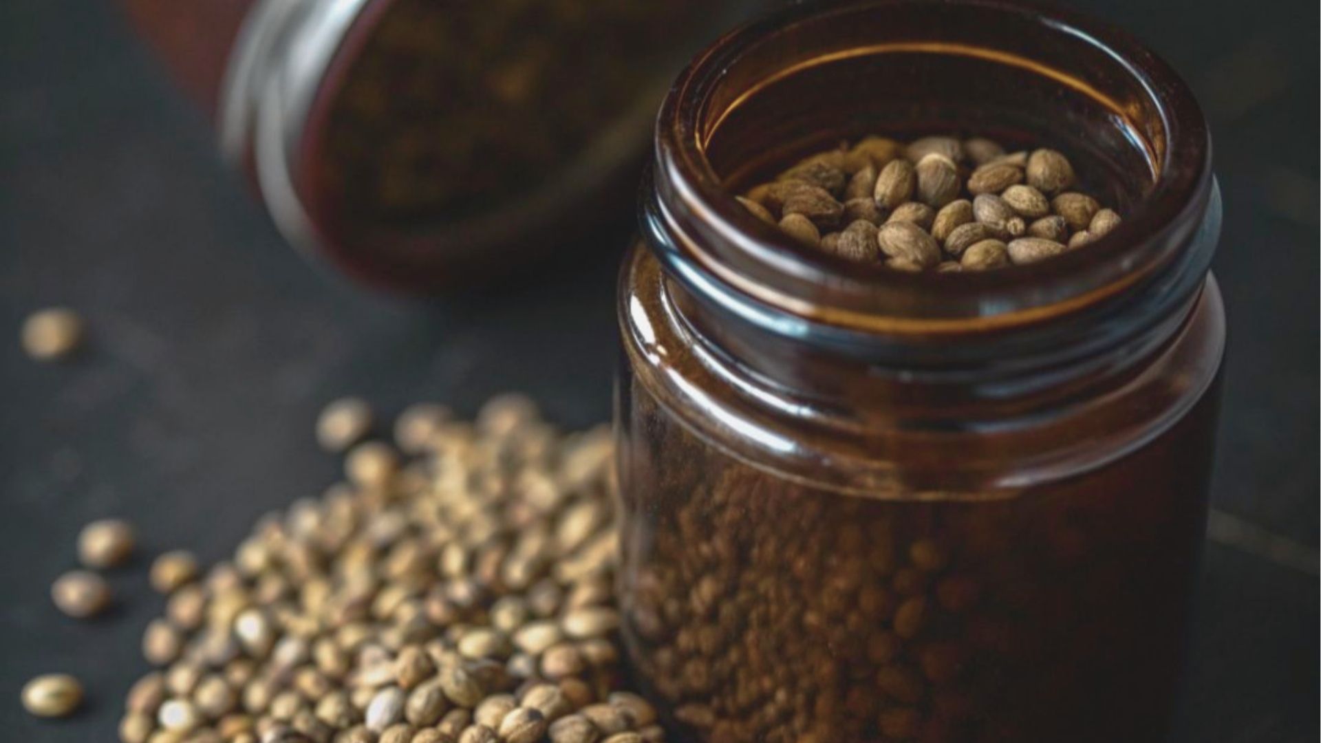 Amber glass jar filled with cannabis seeds, with more seeds scattered around the open lid on a dark background