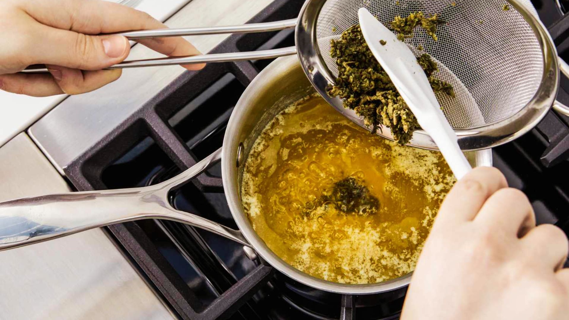 Hands strain decarboxylated cannabis from melted butter using a mesh strainer over a saucepan on the stove.