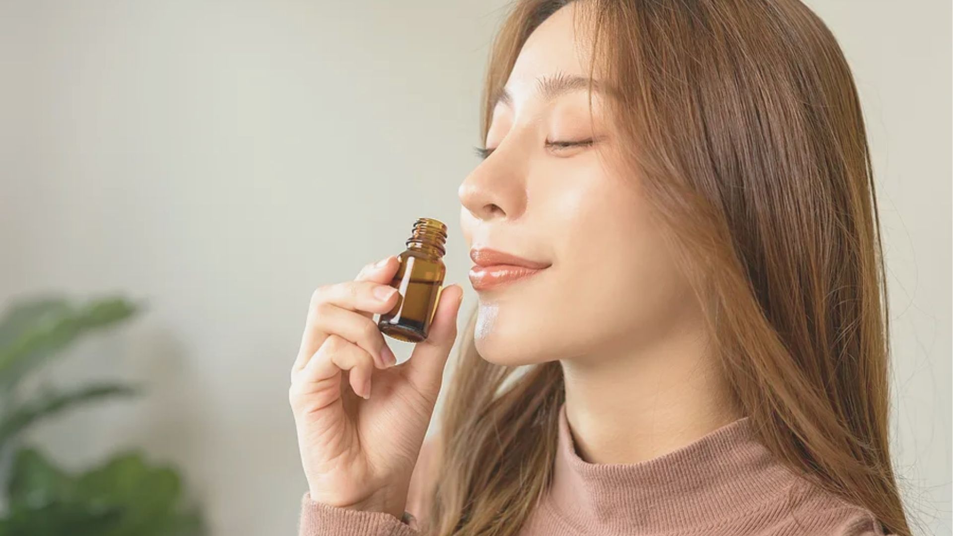 A woman in a pink sweater with eyes closed smiles while holding a small amber CBD oil bottle to her nose.