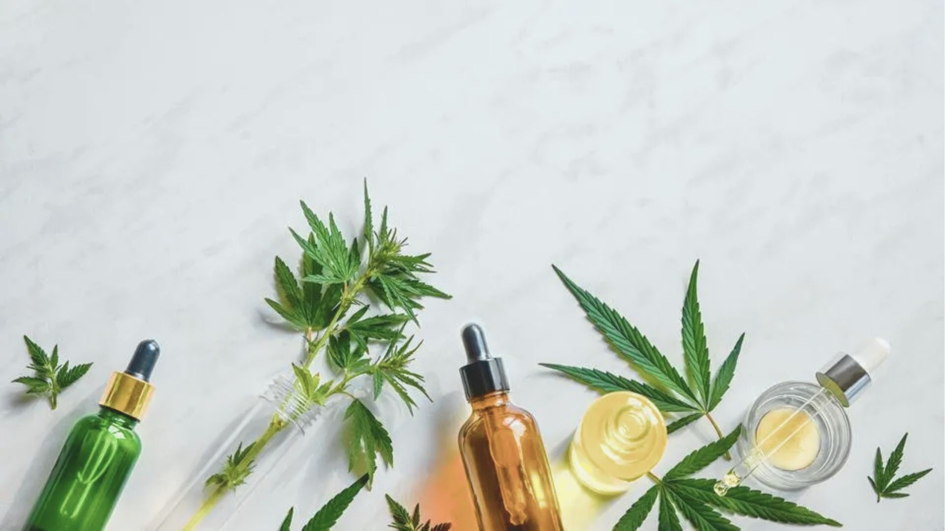 Flat lay of CBD oil dropper bottles in amber and green glass surrounded by hemp leaves on a white marble background.