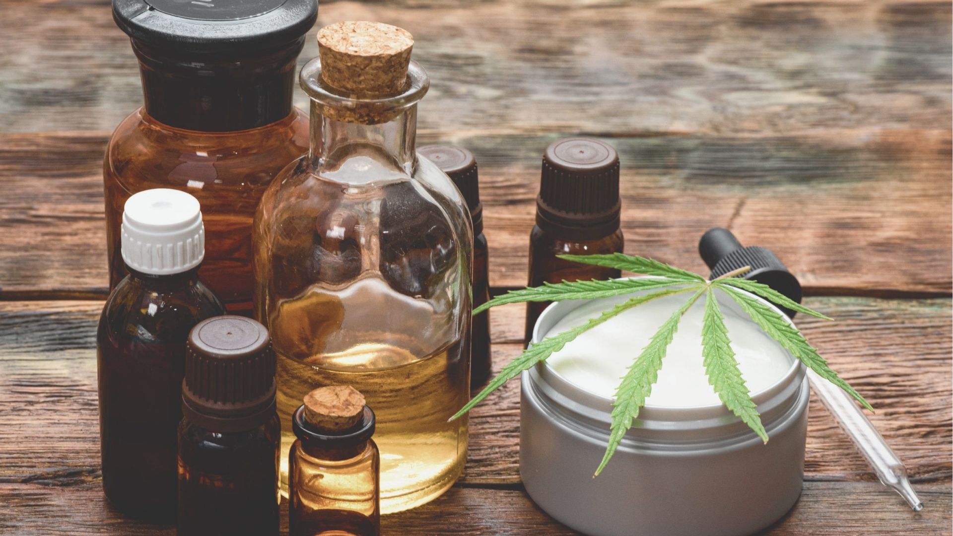 Assortment of amber glass dropper bottles, a corked vial, and a jar of topical cream with a cannabis leaf on a rustic wood table