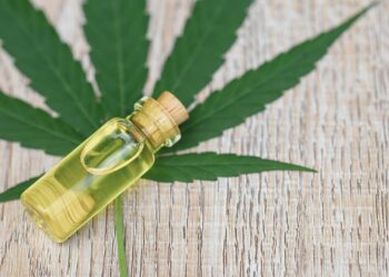 Small corked vial of yellow CBD oil resting on a large cannabis leaf over a wooden surface
