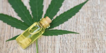 Small corked vial of yellow CBD oil resting on a large cannabis leaf over a wooden surface