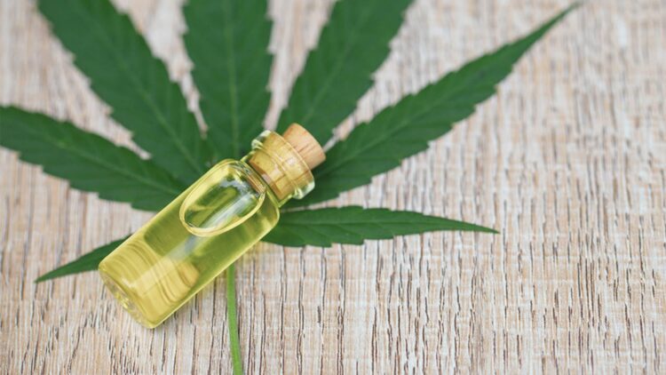 Small corked vial of yellow CBD oil resting on a large cannabis leaf over a wooden surface