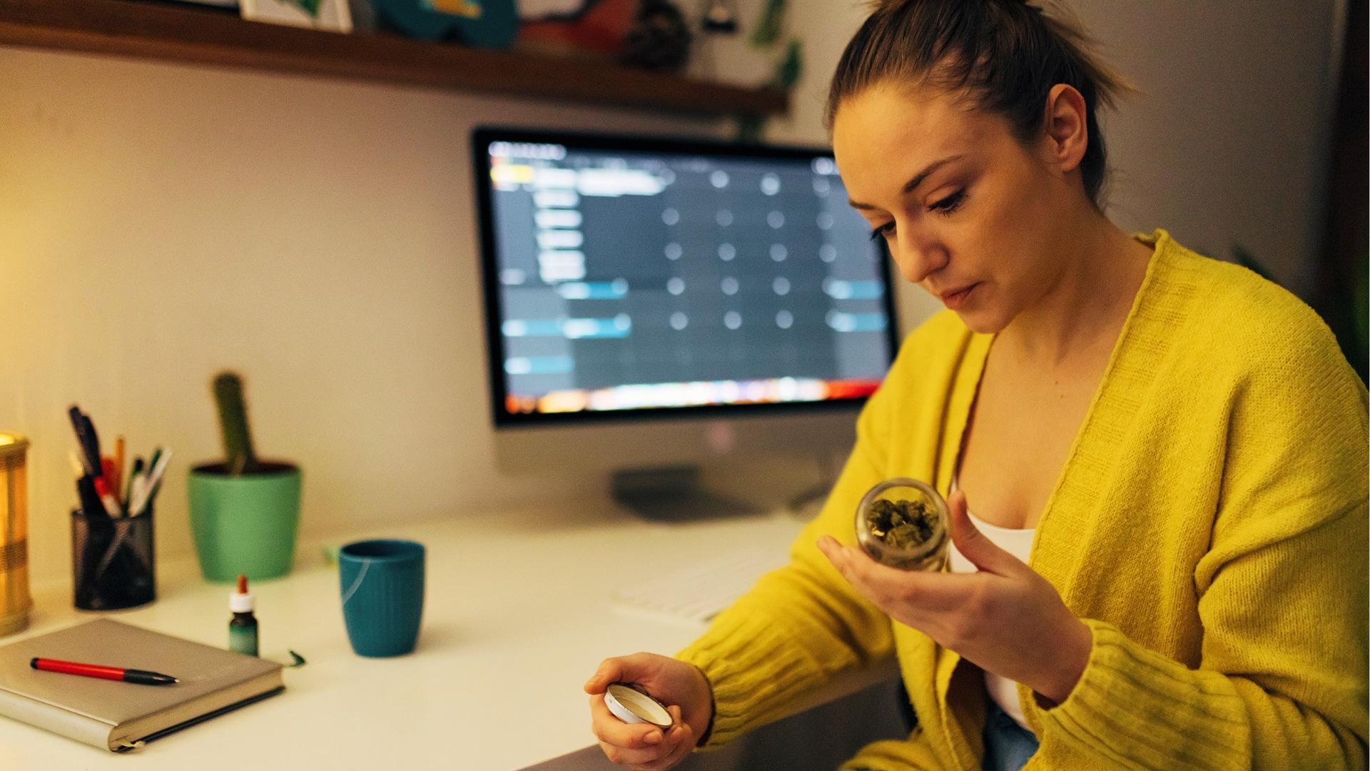 Woman in yellow cardigan holds small glass jar of cannabis buds at desk with computer calendar, mug, and notebook nearby.