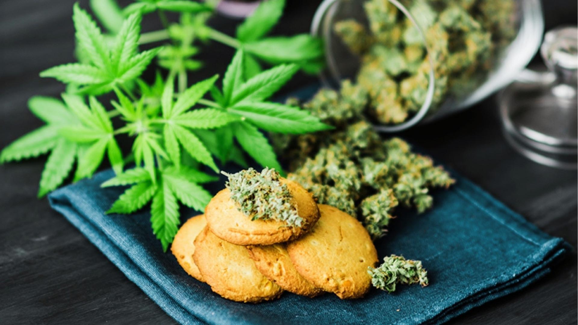 Cannabis-infused cookies topped with green buds sit on blue napkins beside fresh leaves and tipped glass jar spilling flowers.