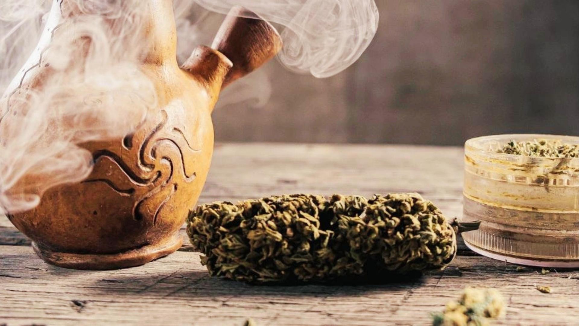 Ornate clay smoking pipe emits wisps of smoke beside a large cannabis bud and plastic grinder filled with ground flowers on wood.