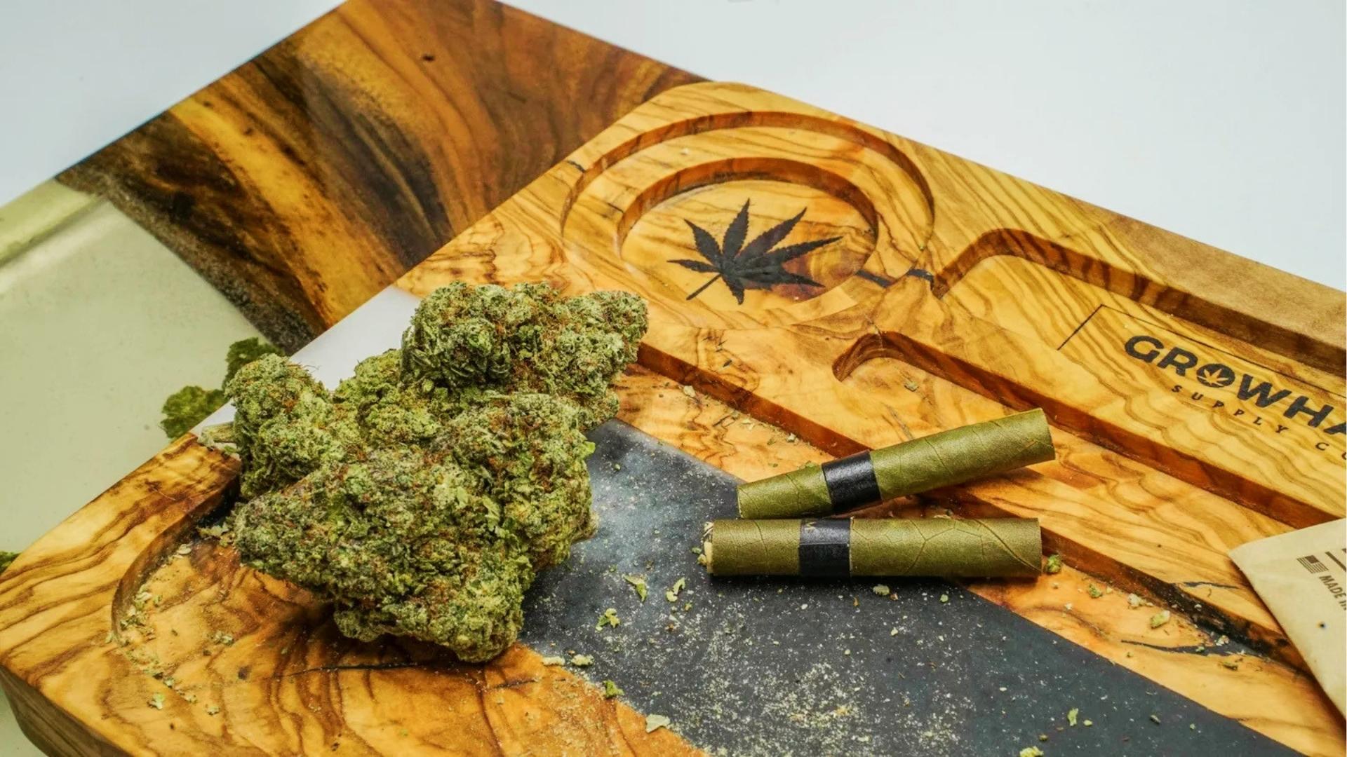 Large cannabis bud rests on a custom olive wood tray with engraved leaf beside two green pre-rolled blunts and rolling papers.
