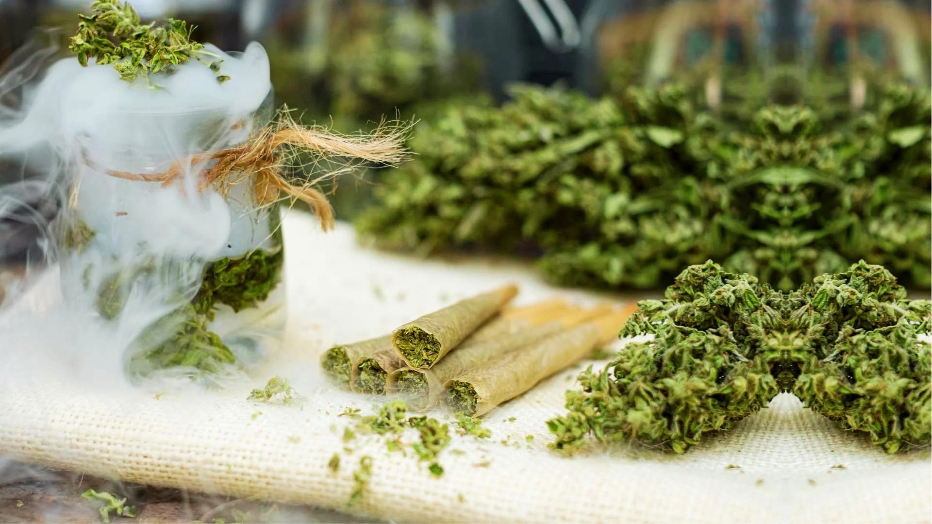 Glass jar wrapped in twine with dry ice smoke and cannabis buds sits beside pre-rolled joints and loose flowers on burlap cloth.