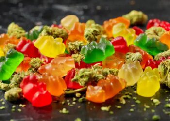 Assortment of colorful cannabis gummies and brownies displayed beside frosty green buds and ground flowers on dark stone surface.