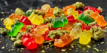Assortment of colorful cannabis gummies and brownies displayed beside frosty green buds and ground flowers on dark stone surface.
