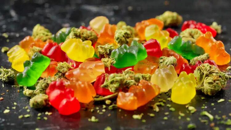 Assortment of colorful cannabis gummies and brownies displayed beside frosty green buds and ground flowers on dark stone surface.