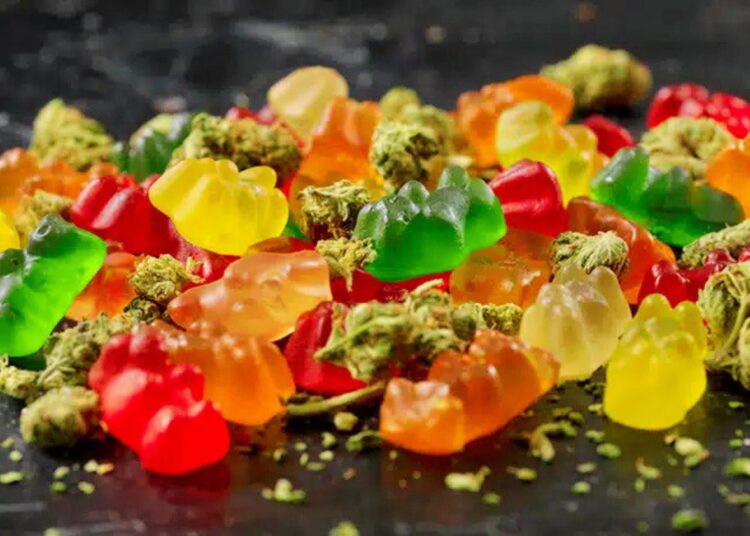 Assortment of colorful cannabis gummies and brownies displayed beside frosty green buds and ground flowers on dark stone surface.